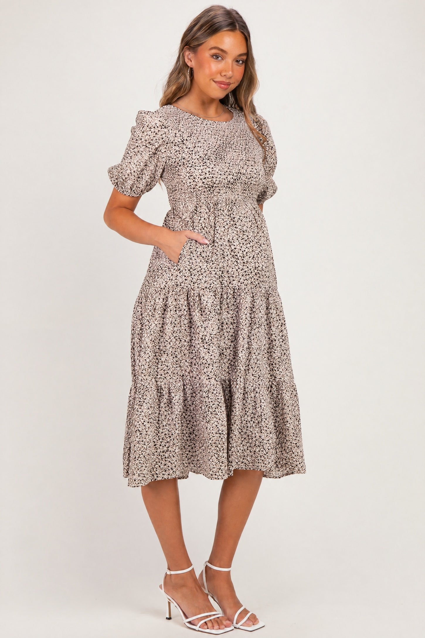woman wearing a beige floral print maternity dress with short puff sleeves and pockets, standing with a baby bump visible