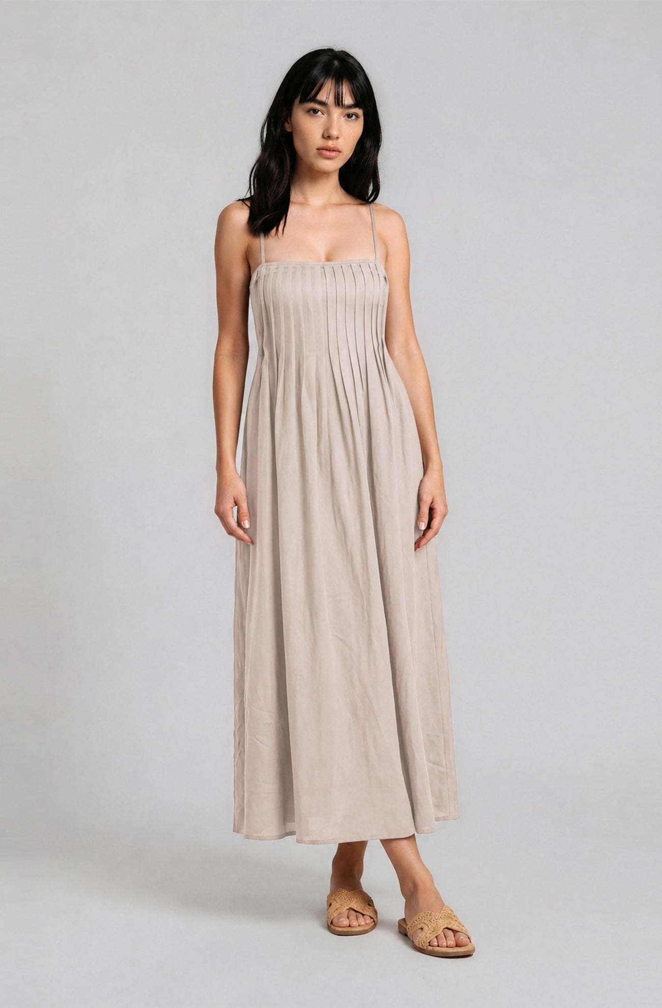 woman wearing a beige spaghetti strap maxi dress with pleated details and standing in a neutral background