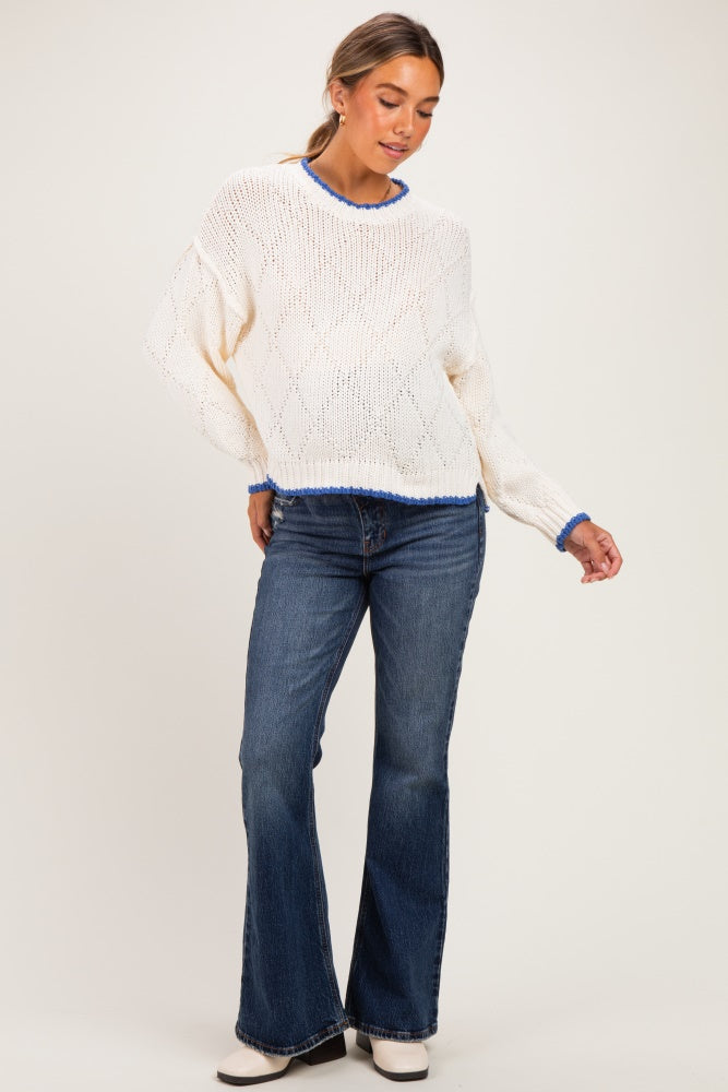 woman wearing a white laced maternity sweater with blue trim and blue flared jeans while standing and posing