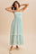 woman wearing a light green floral maxi dress with adjustable straps and ruffled hem, posing with hands on her head