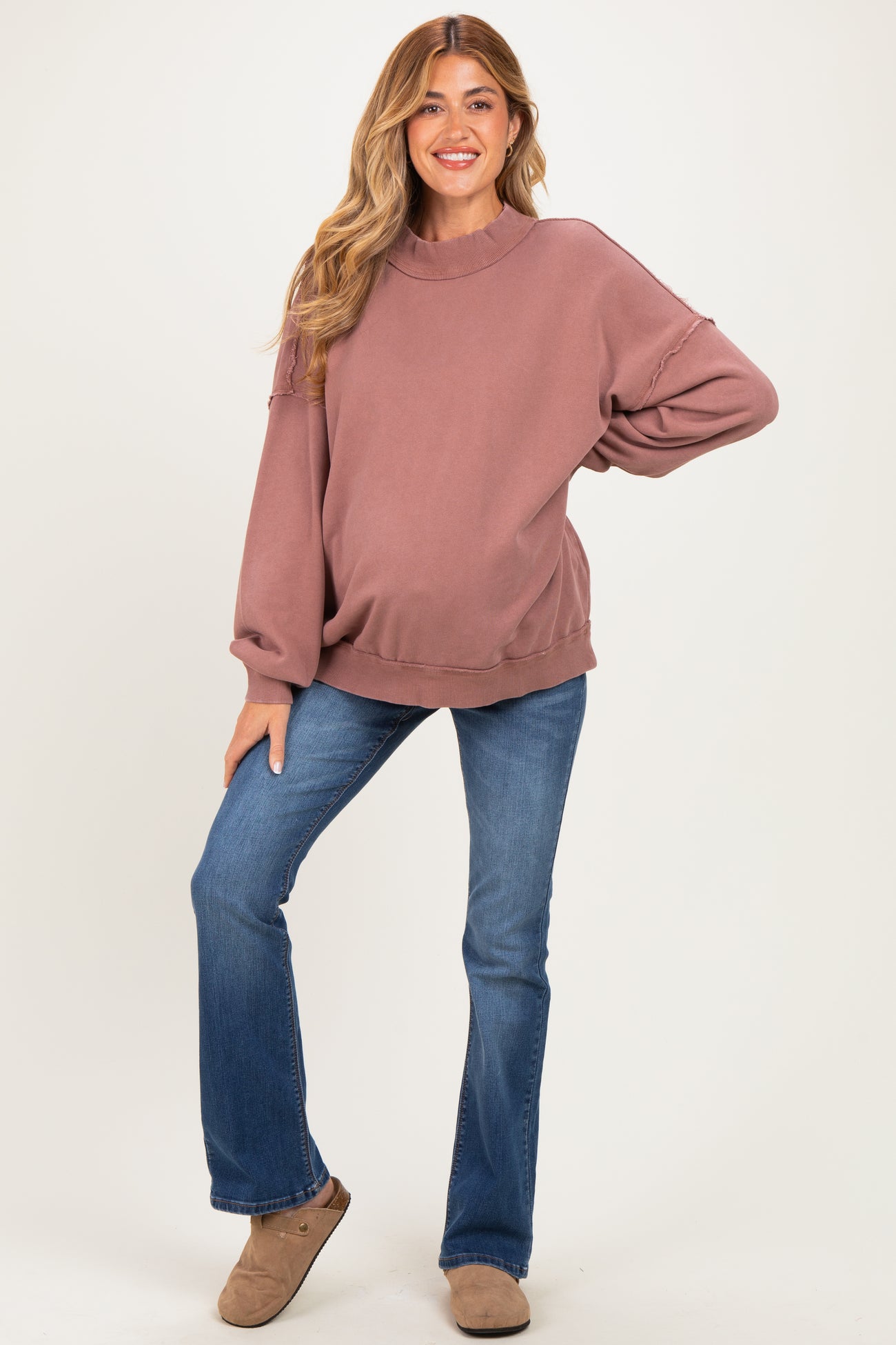 woman wearing a dusty rose oversized maternity sweatshirt and blue flared jeans, with a visible baby bump
