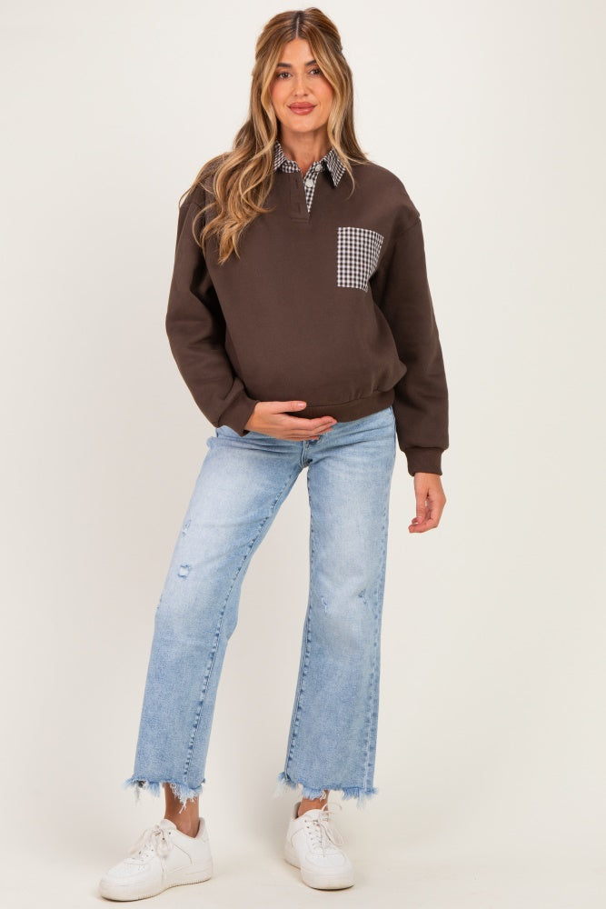 woman wearing a brown maternity sweatshirt with a checked pocket and blue distressed jeans, holding her baby bump