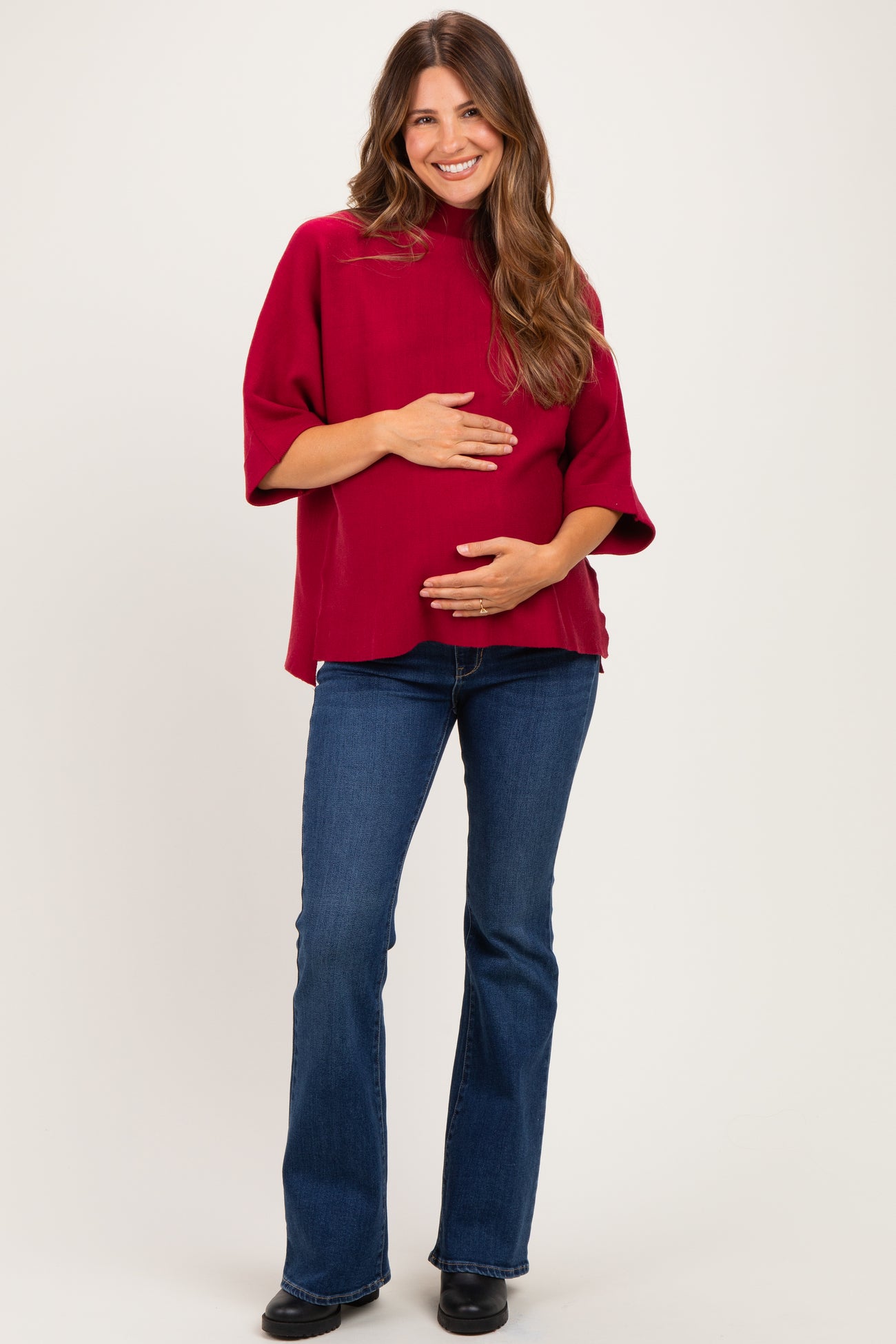 woman wearing a burgundy maternity top and flared jeans, holding her baby bump