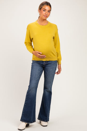 woman wearing a mustard yellow maternity sweater and flared jeans, holding her baby bump