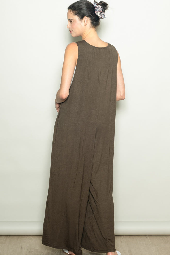 woman wearing a long brown sleeveless maxi dress, standing with her back to the camera and hands in pockets
