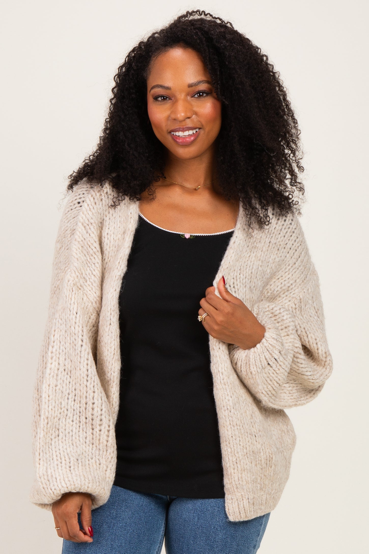 woman wearing a chunky beige knit cardigan over a black tank top and blue jeans, smiling at the camera