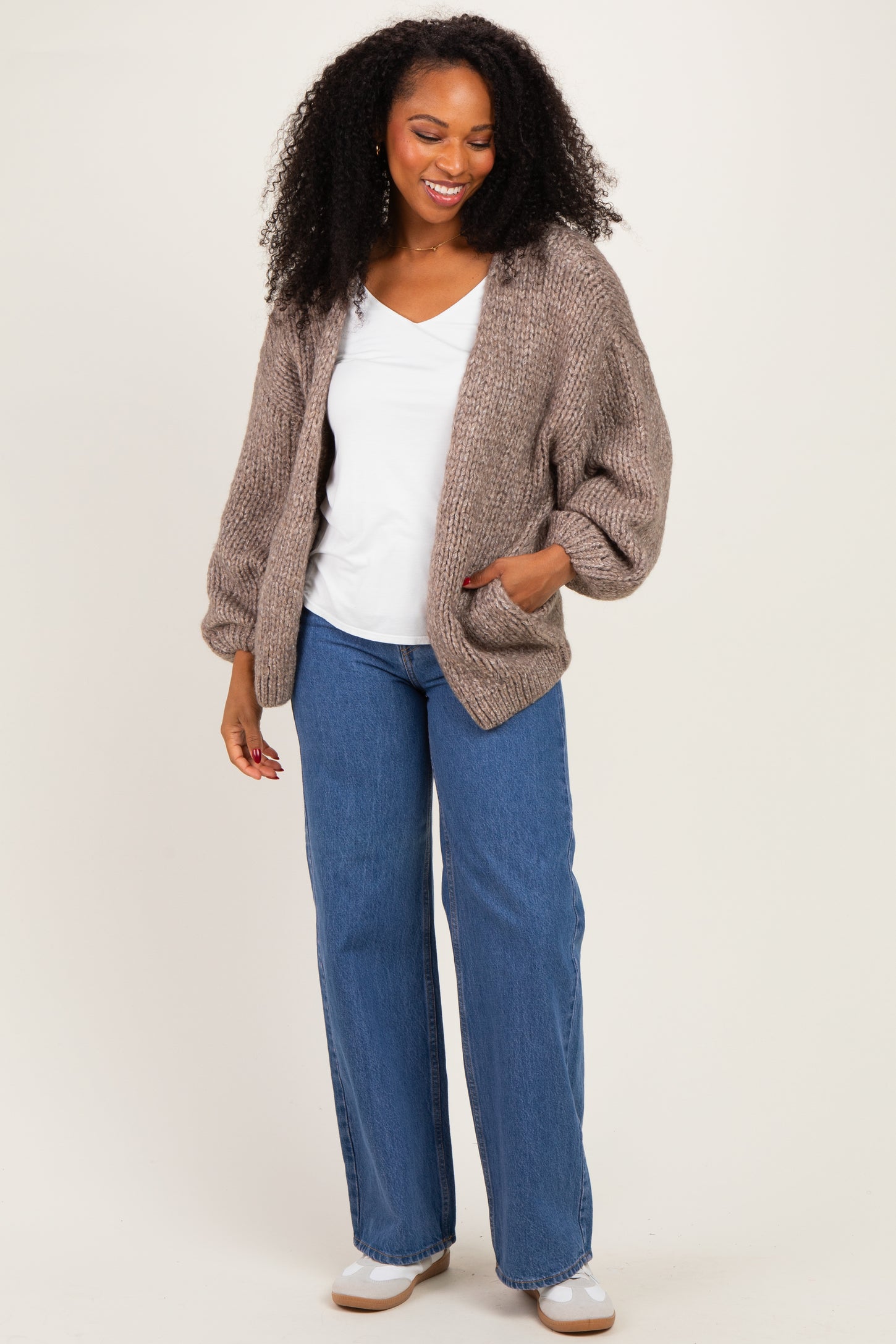 woman wearing a beige oversized knitted cardigan with pockets, white v-neck top, and wide-leg blue jeans
