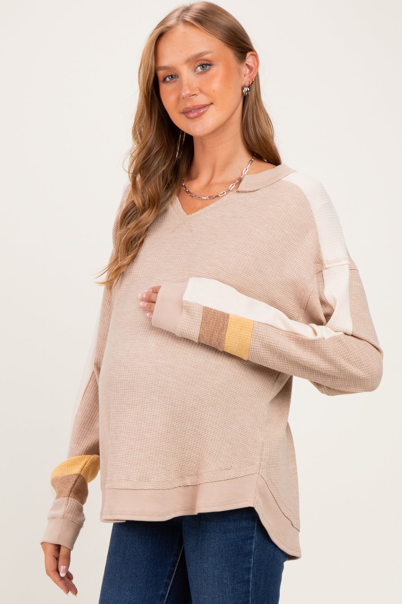woman wearing a beige v-neck maternity sweater with color block sleeves and holding her baby bump