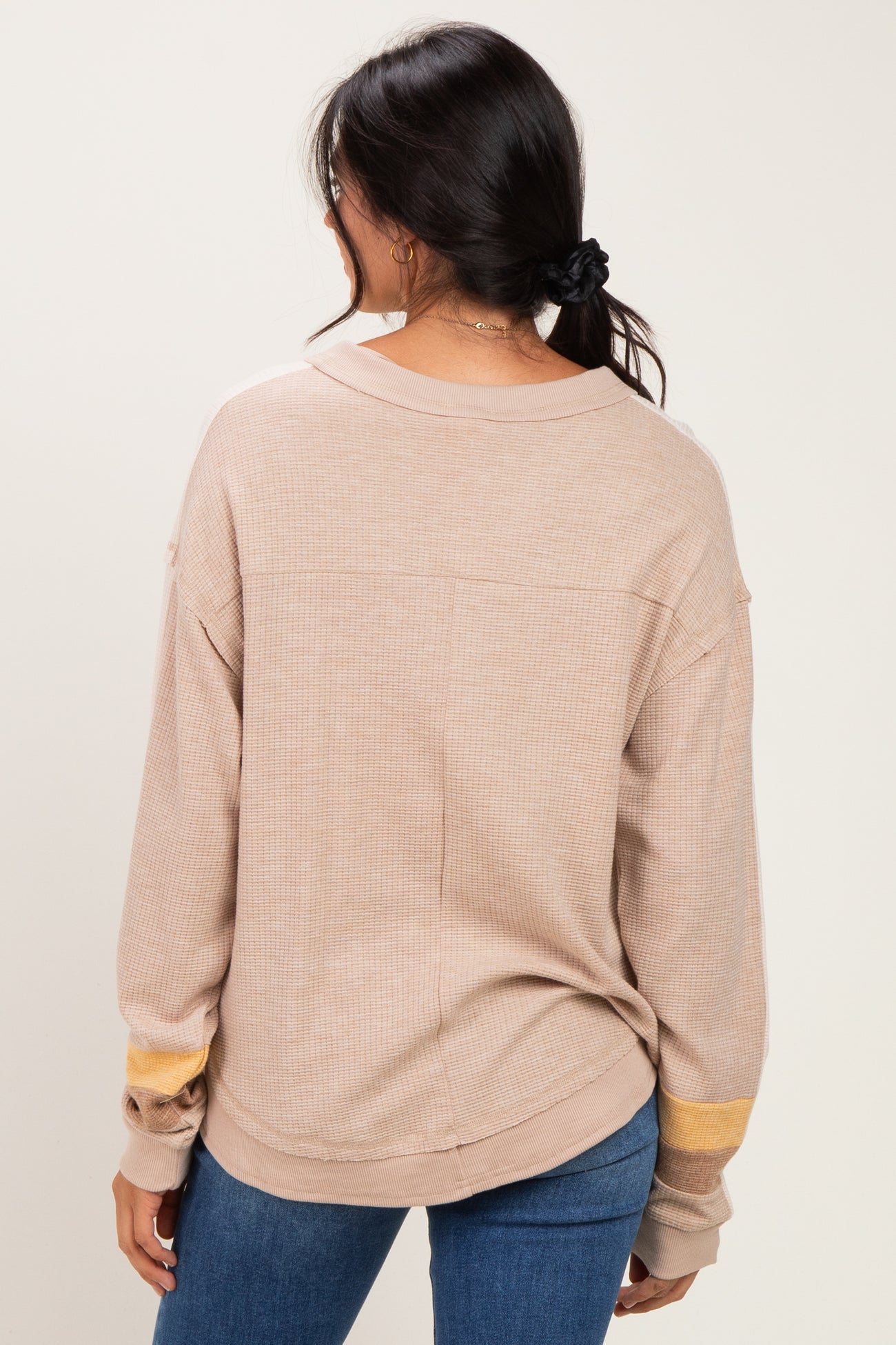 woman wearing a tan oversized long sleeve top with yellow stripe detail on the sleeves, standing backwards