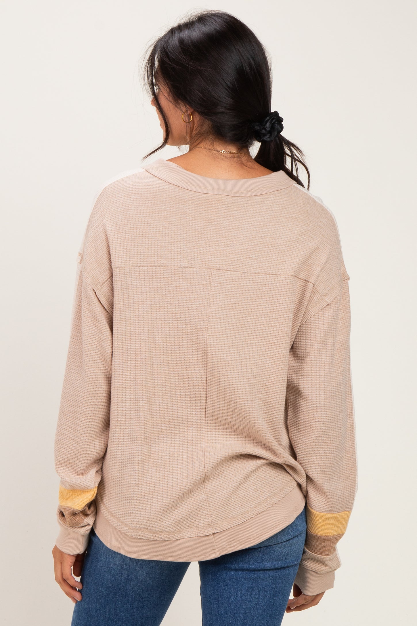 woman wearing a tan oversized long sleeve top with yellow stripe detail on the sleeves, standing backwards