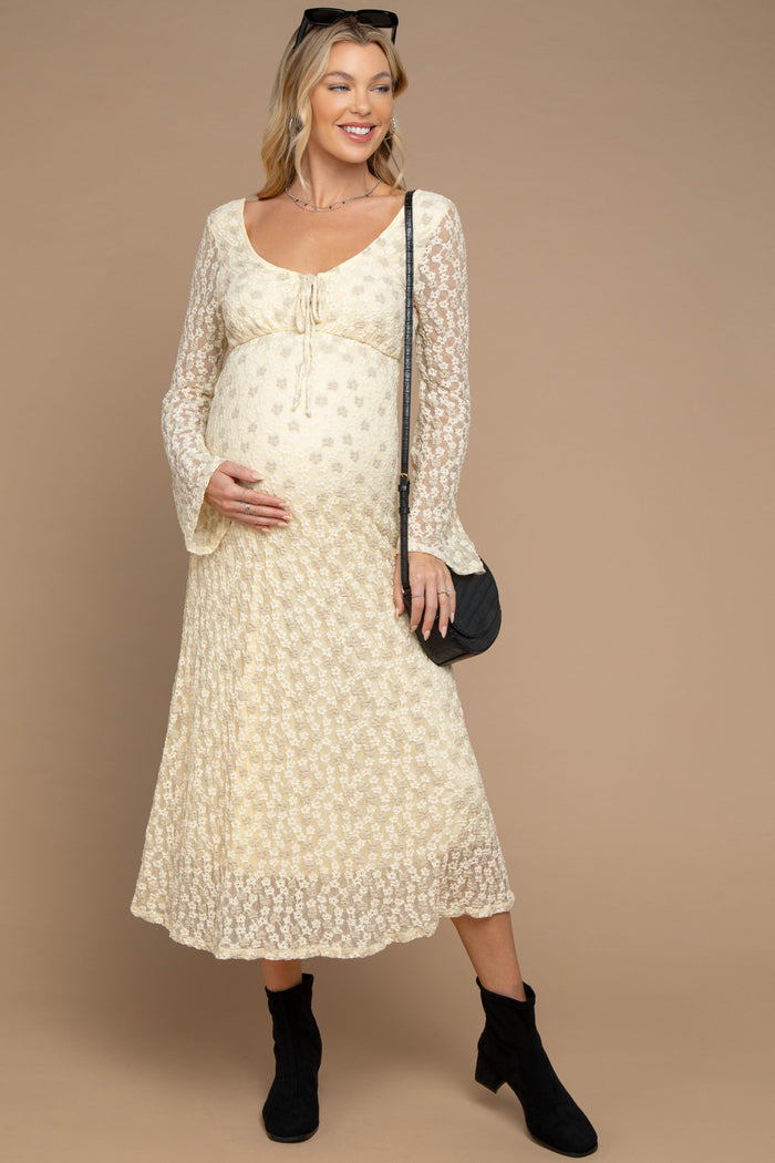 woman wearing a cream floral lace maternity dress with a baby bump and bell sleeves, holding her belly with a black purse and boots
