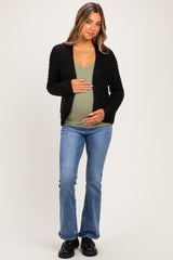 woman wearing a black knitted maternity cardigan over a green tank top and holding her baby bump, paired with flared jeans