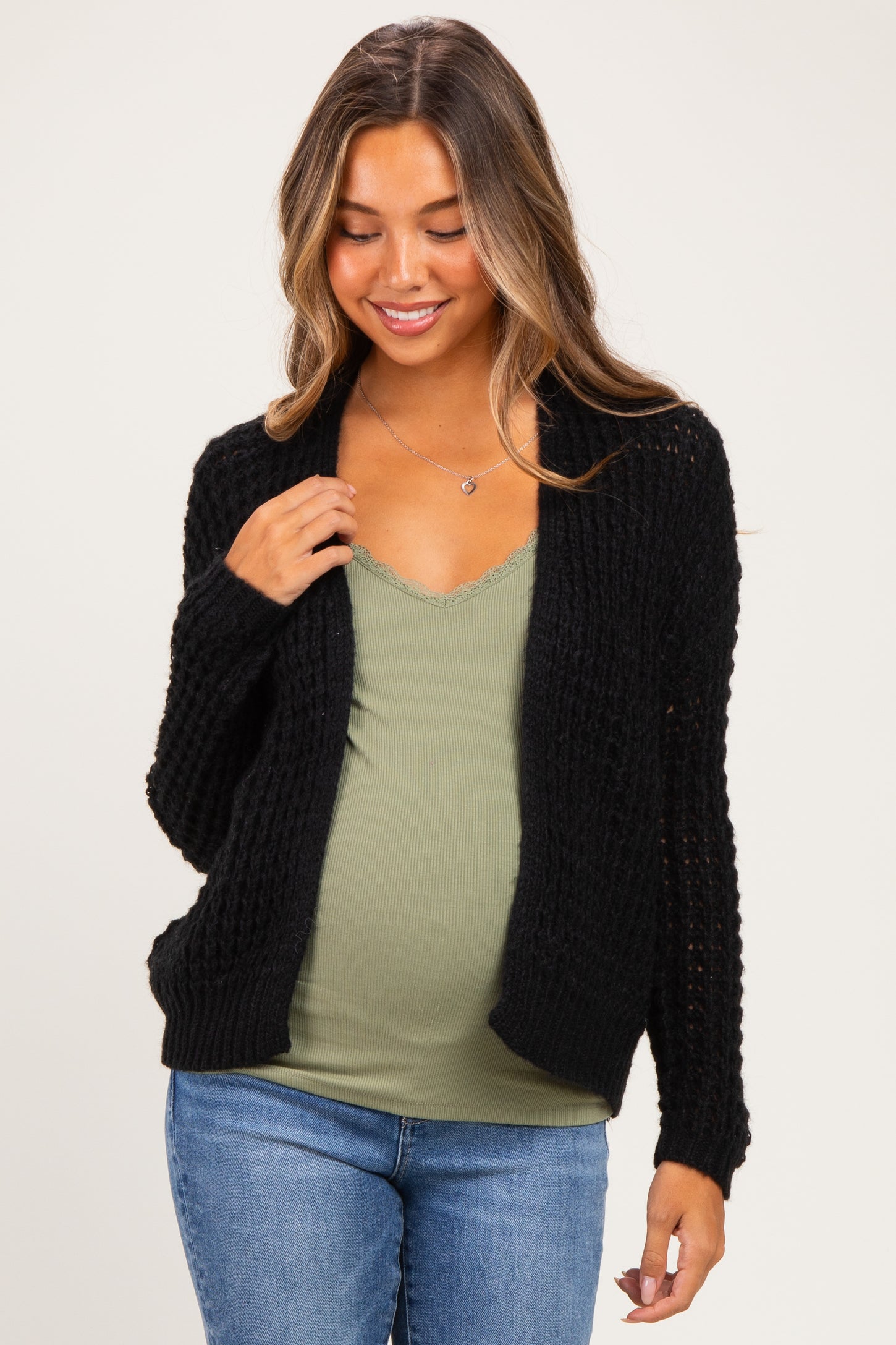 woman wearing a black chunky knit maternity cardigan over a green tank top and showing her baby bump, paired with blue jeans