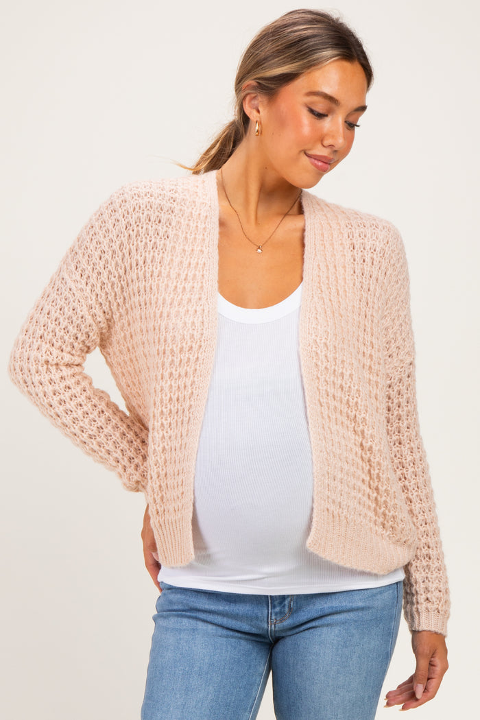 woman wearing a light pink open-knit maternity cardigan over a white tank top, showcasing her baby bump