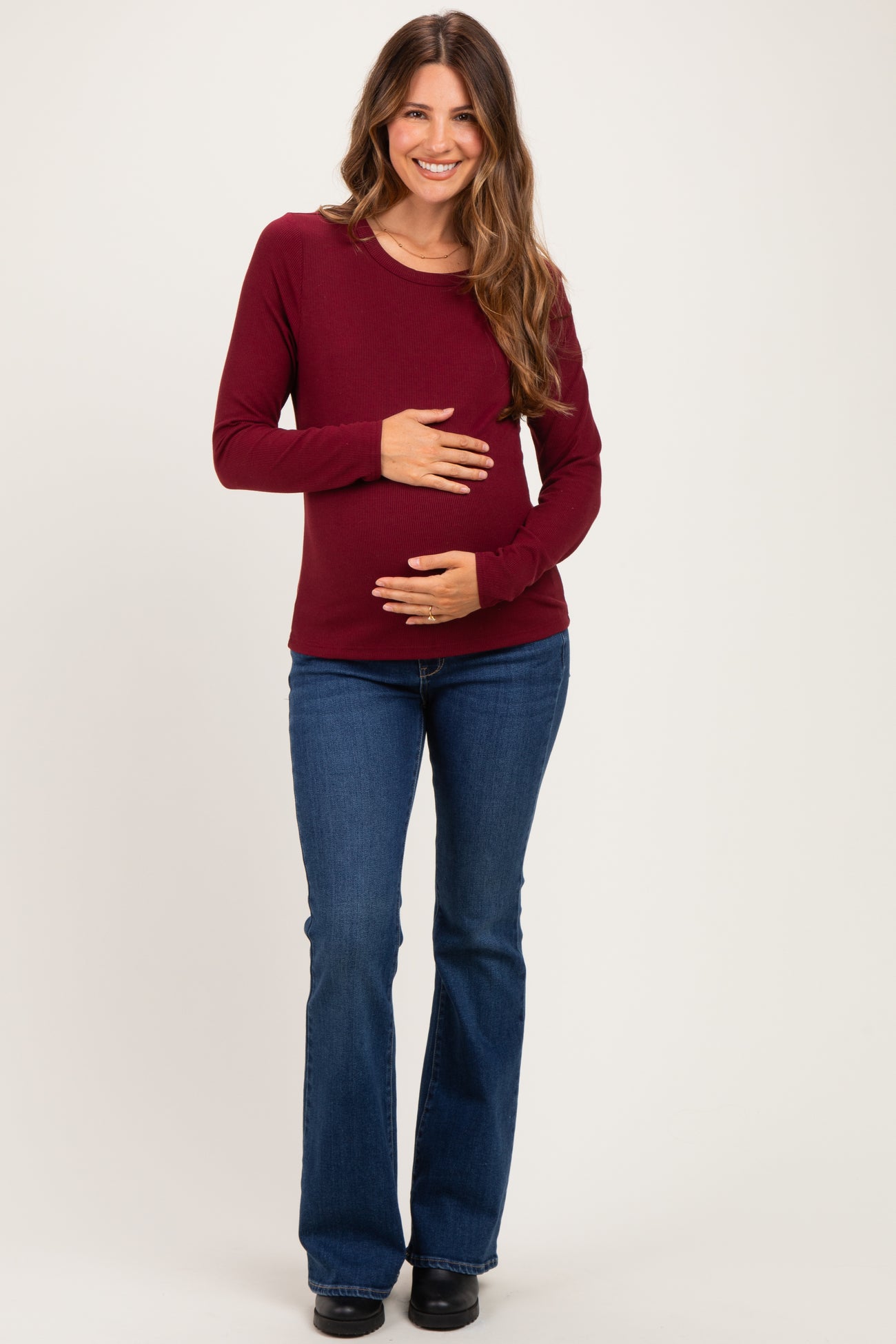 woman wearing a burgundy long sleeve maternity top and blue flared jeans, gently cradling her baby bump