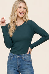 woman wearing a dark green ribbed long sleeve top and blue jeans, smiling with one hand raised