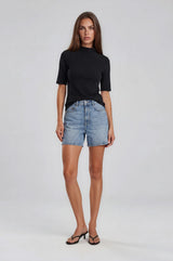 woman wearing a black short sleeve top and light blue denim shorts, standing against a neutral background