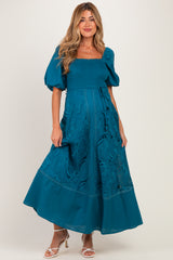 woman wearing a teal maternity maxi dress with puff sleeves and lace detailing, showcasing her baby bump