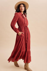 woman wearing a rust colored long sleeve button-up maxi dress with an elastic waist and ruffle hem, paired with a wide-brim hat and boots
