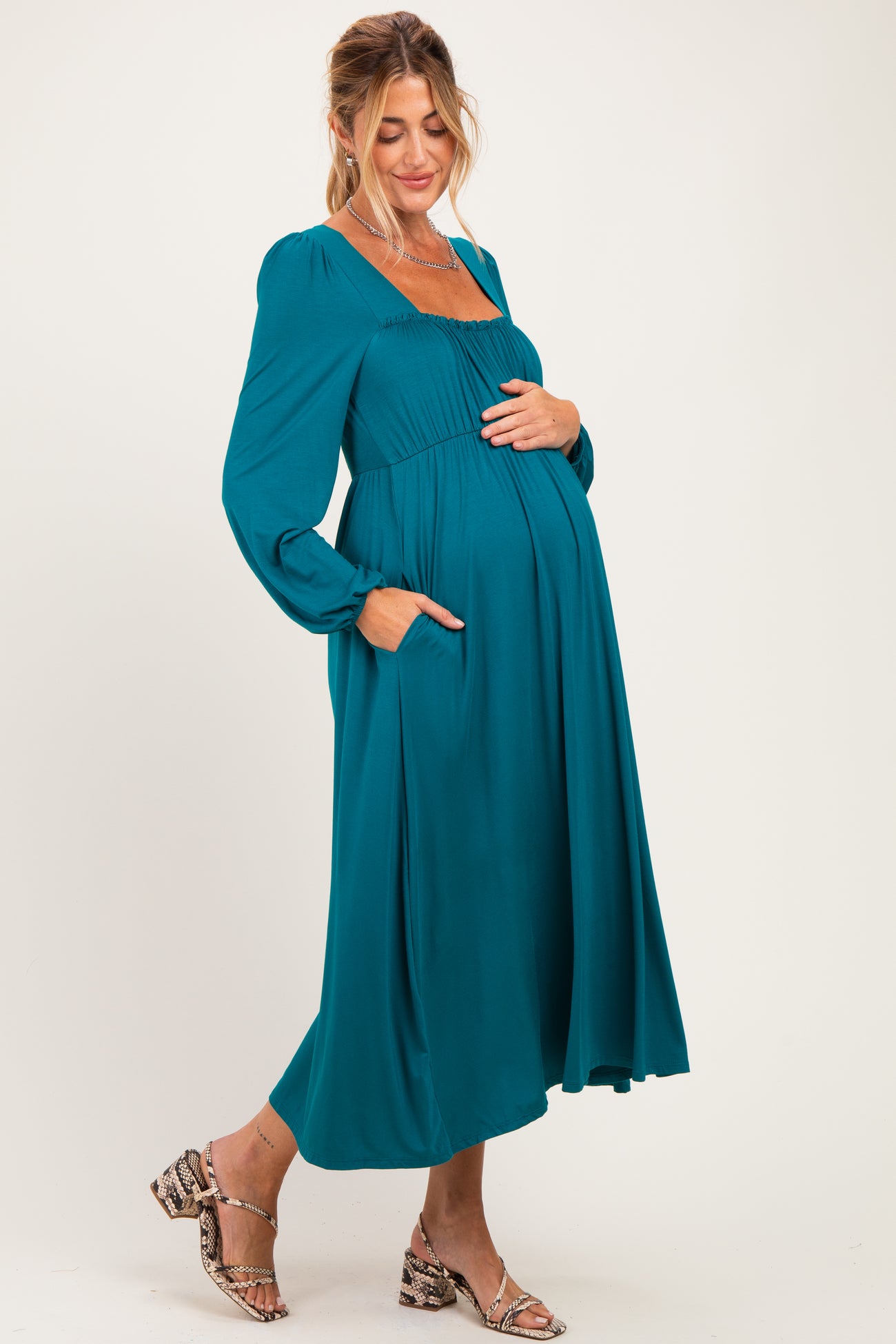 woman wearing a teal long sleeve maternity dress with a square neckline and pockets, showing off her baby bump
