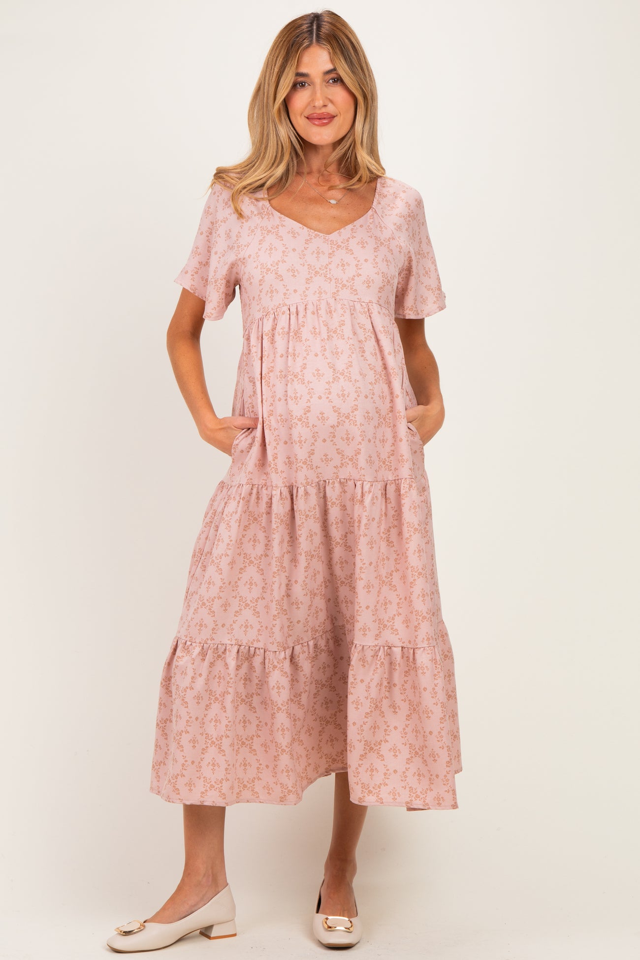 woman wearing a light pink maternity dress with a v-neck, short sleeves, and tiered skirt, holding her baby bump