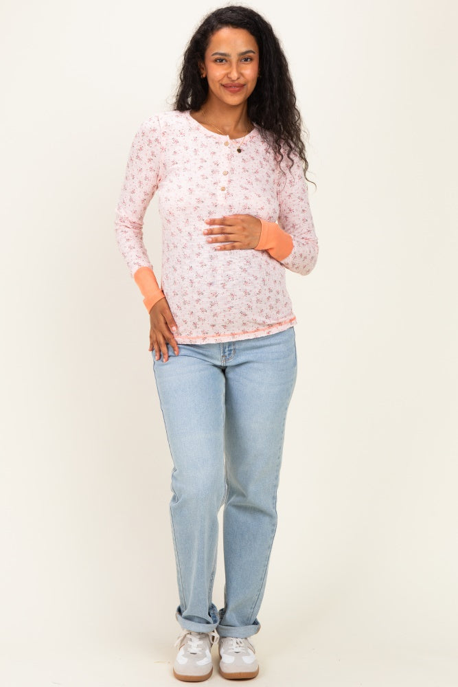 woman wearing a light pink floral maternity top with long sleeves and buttons, holding her baby bump and paired with light blue jeans