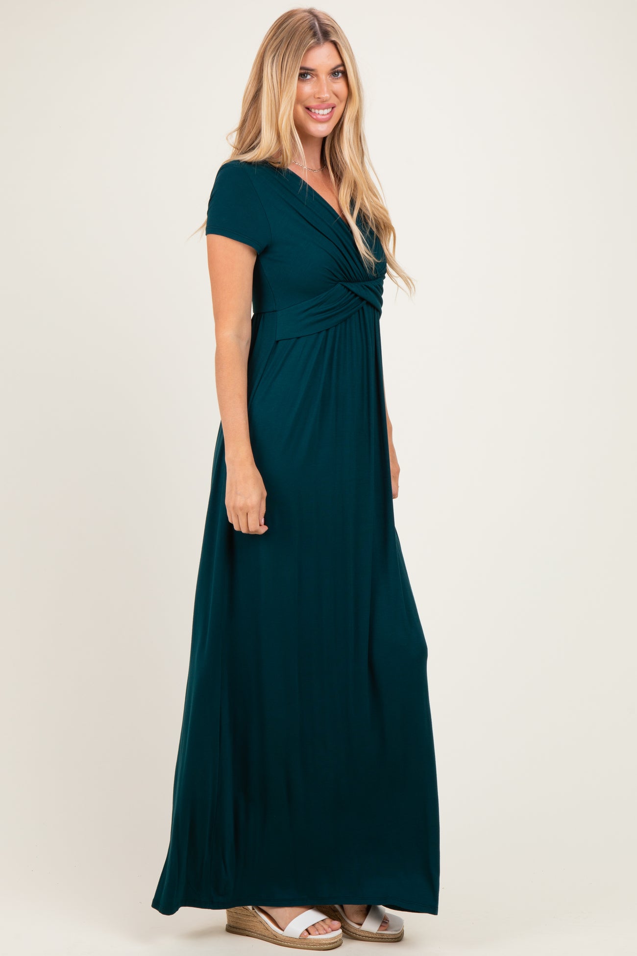 woman wearing a teal maxi dress with short sleeves and a gathered bodice, standing sideways with a neutral background
