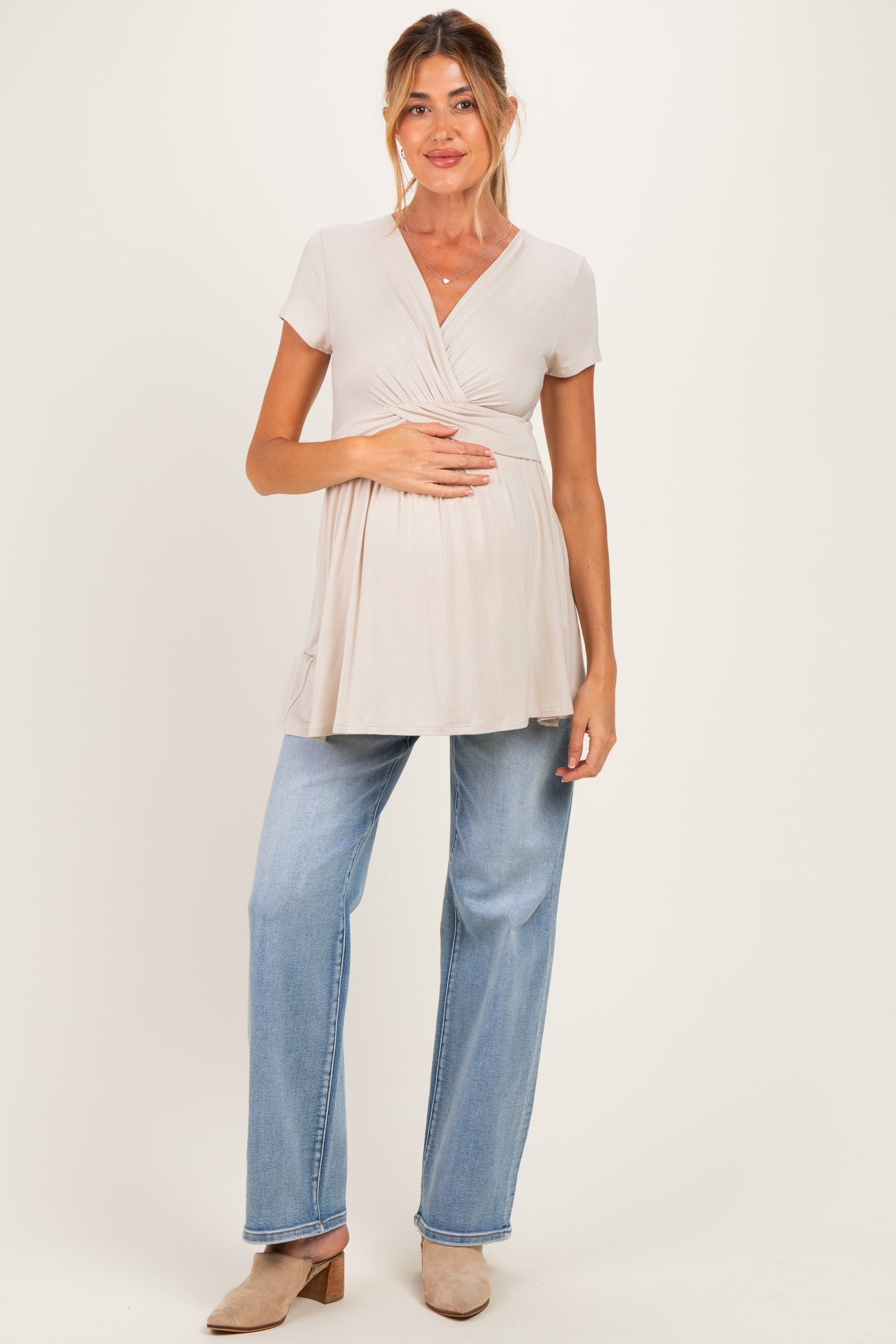woman wearing a light beige maternity top with a wrap design and a visible baby bump, paired with blue jeans and neutral shoes