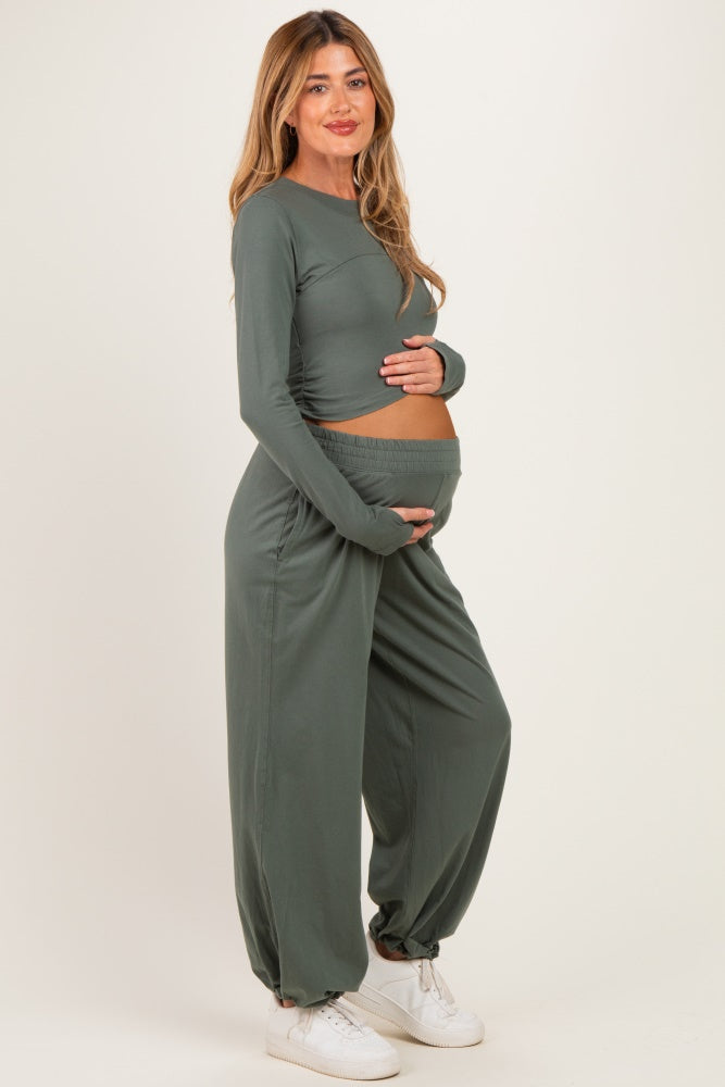 woman wearing a sage green long sleeve maternity top and matching pants, cradling her baby bump