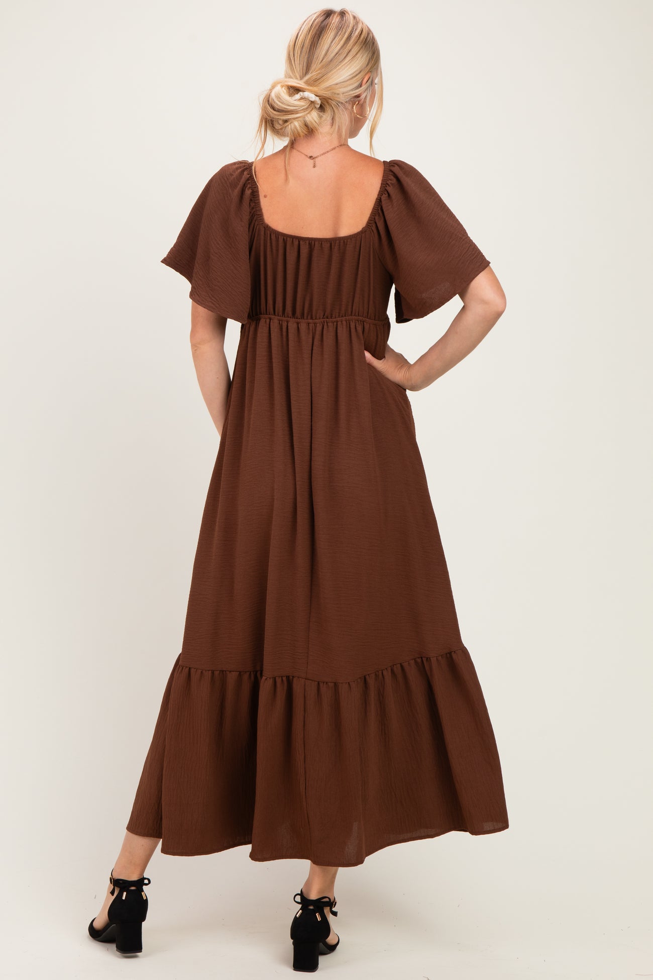 woman wearing a brown maxi dress with short flutter sleeves and a tiered hem, standing with hands in pockets