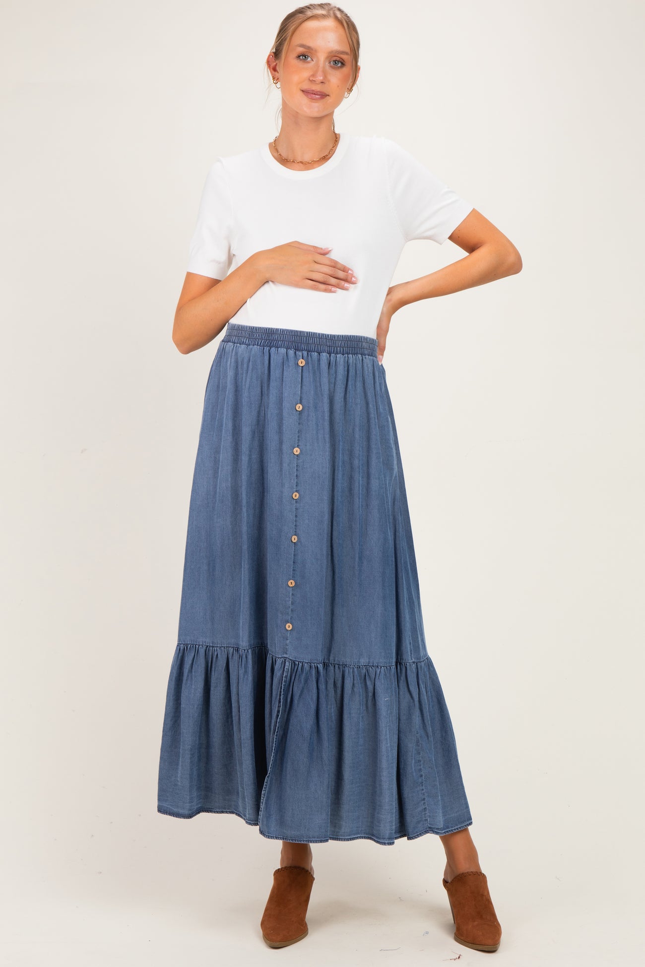 woman wearing a white short sleeve top and a blue maternity maxi skirt with buttons, holding her baby bump