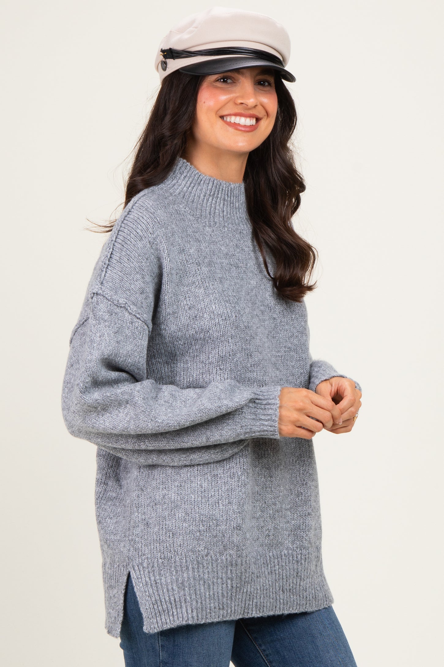 woman wearing a grey oversized sweater with a high neckline and side slits, paired with a stylish cap