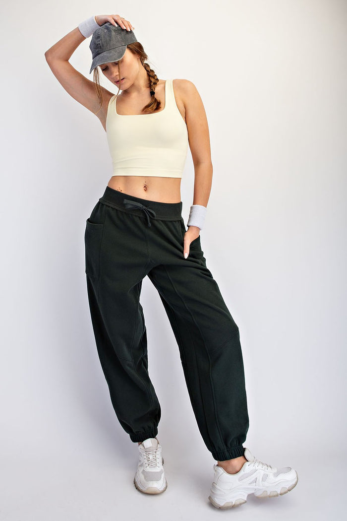 woman wearing a light yellow crop top and dark green sweatpants with pockets, styled with a cap and white sneakers