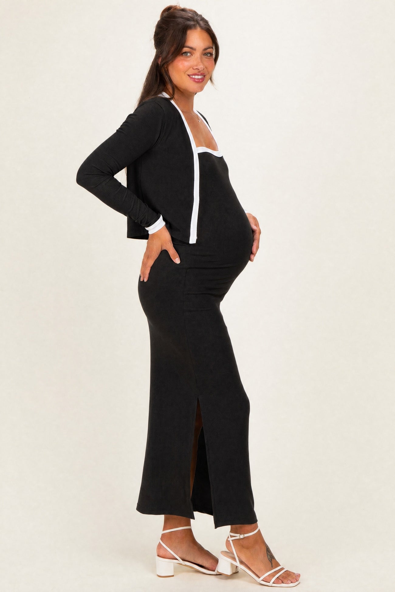 woman wearing a black maternity dress with a side slit and a matching open cardigan, gently cradling her baby bump