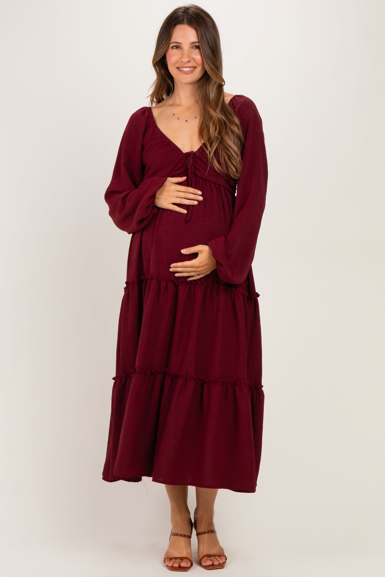 woman wearing a burgundy maternity maxi dress with long sleeves and tiered ruffle details while holding her baby bump