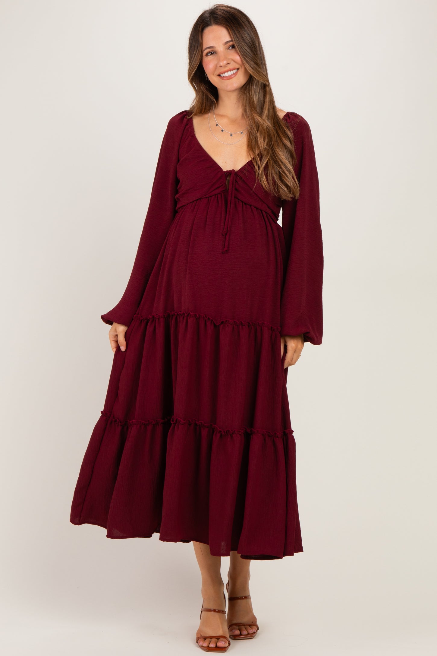 woman wearing a burgundy long sleeve maternity dress with a tiered skirt and a tie detail at the bust, showing her baby bump