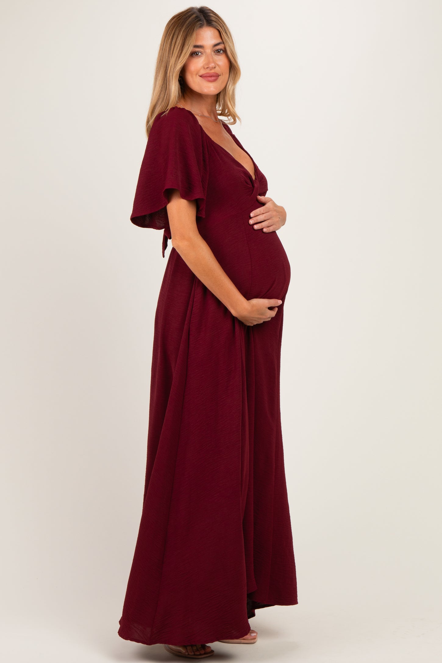 woman wearing a burgundy maternity maxi dress with flutter sleeves and holding her baby bump