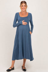 woman wearing a blue long sleeve maternity dress with a scoop neckline and pockets, cradling her baby bump