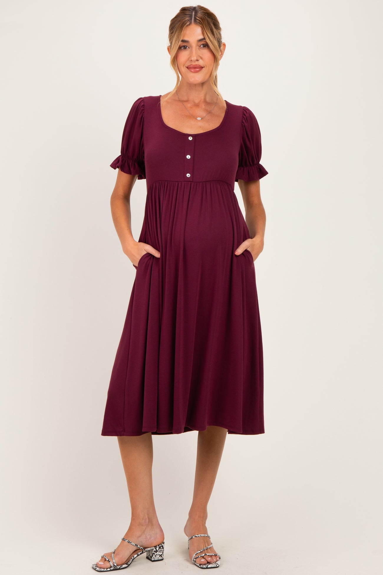 woman wearing a burgundy maternity dress with short puff sleeves, pockets, and buttons down the front, showcasing her baby bump