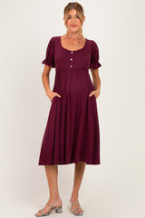 woman wearing a burgundy maternity dress with short puff sleeves, pockets, and buttons down the front, showcasing her baby bump