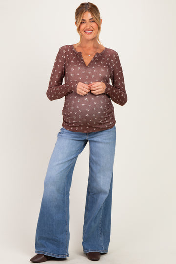 woman wearing a brown maternity top with a v-neck and floral print, paired with light blue flared jeans and showing her baby bump
