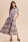woman wearing a long plaid dress with short sleeves, button-down front, and a tied waist, standing against a beige background
