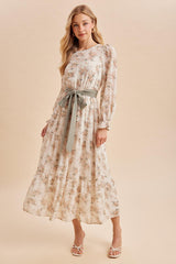 woman wearing a floral midi dress with long sleeves and a tied waistbelt, standing against a neutral background