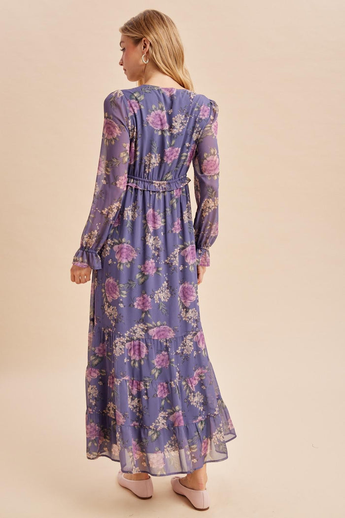 woman wearing a long sleeve purple floral maxi dress with a ruffled hem and waist detail, viewed from the back