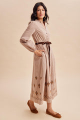 woman wearing a beige button-up midi dress with floral embroidery and a tie belt, paired with brown flats