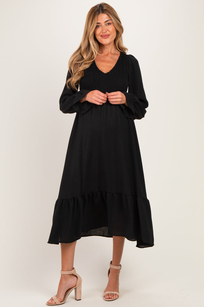 woman wearing a black maternity dress with a smocked bodice and long sleeves, standing in a relaxed pose