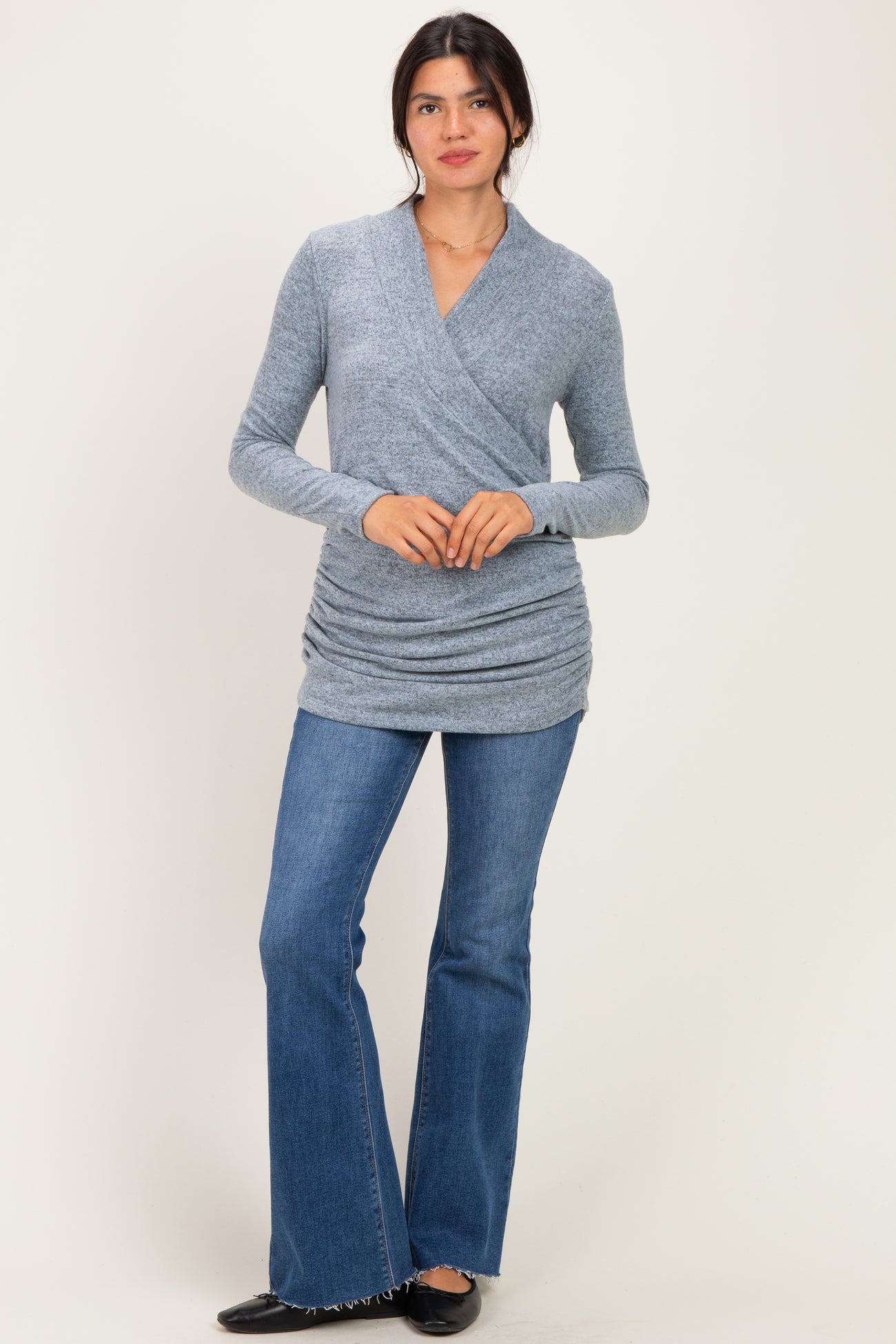 woman wearing a gray wrap-style long sleeve top and flared jeans, standing against a light background