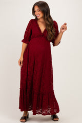 woman wearing a burgundy lace maternity maxi dress with flutter sleeves and a ruffled hem, gently cradling her baby bump