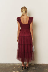 woman wearing a burgundy tiered maxi dress with ruffle sleeves and an open back, standing against a neutral wall