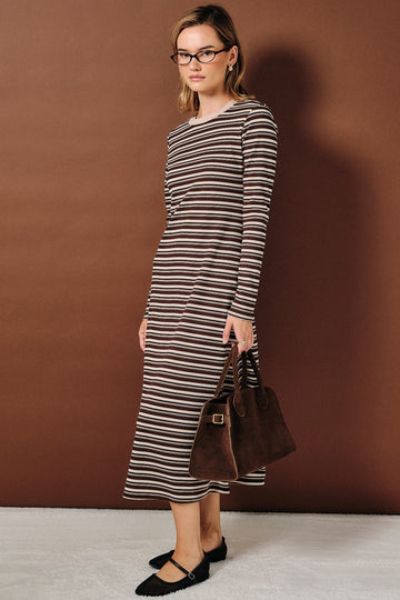 woman wearing a brown and cream striped long sleeve dress, holding a brown handbag, standing against a brown background